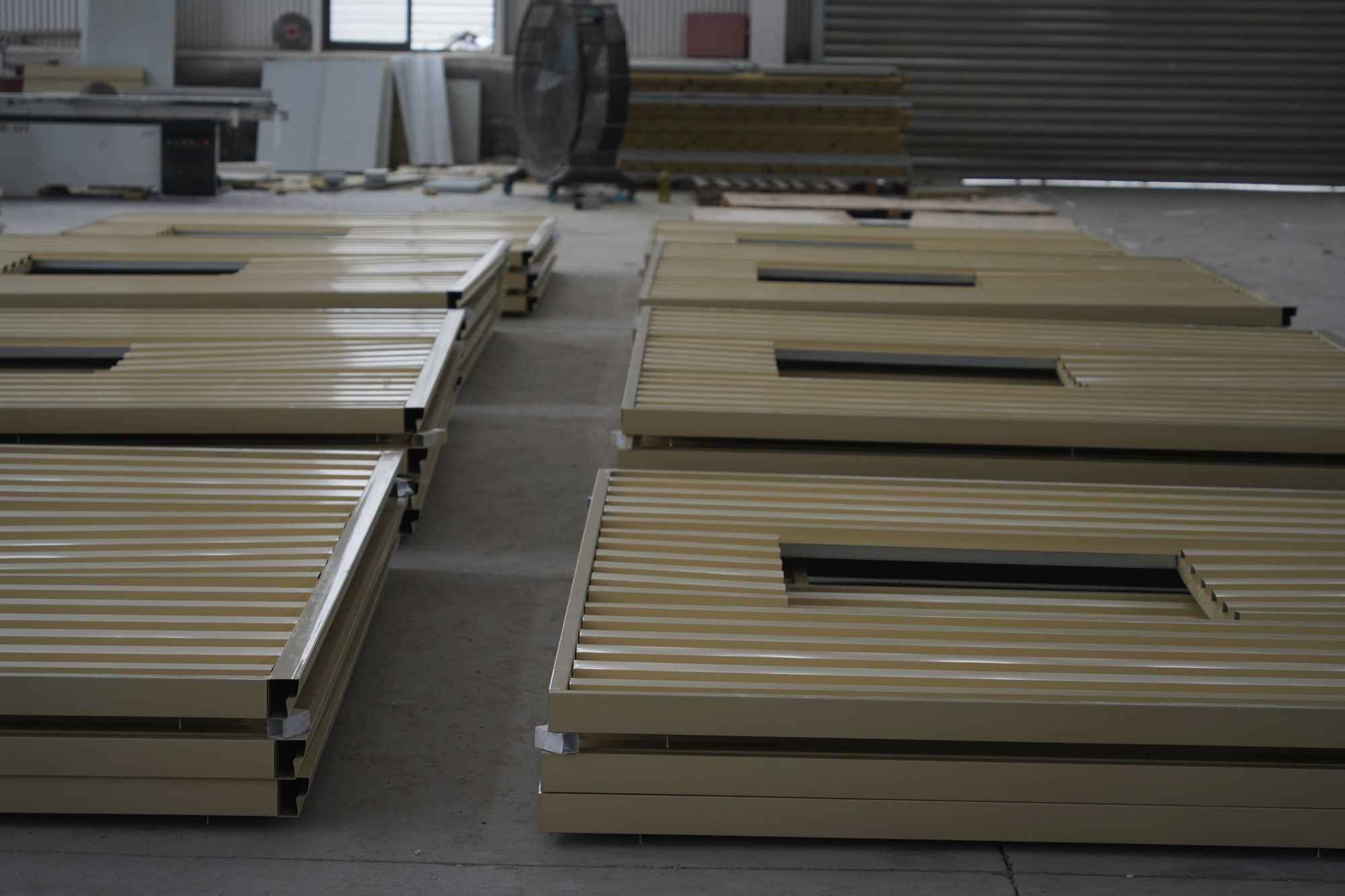 Wall panels stacked flat on a workshop floor, window openings already cut, with a panel saw and more stock behind them.