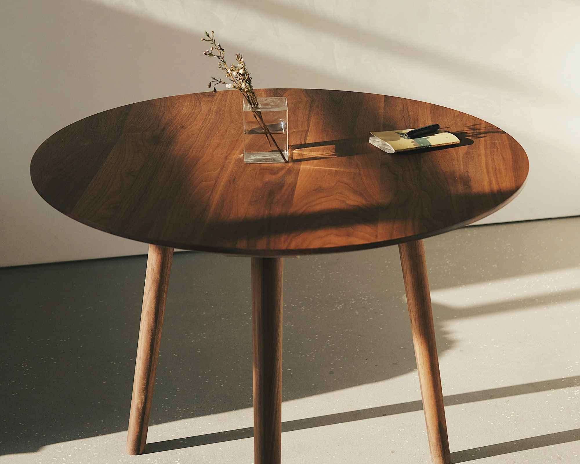 A round solid walnut table on three tapered legs, lit by hard afternoon light from a side window.