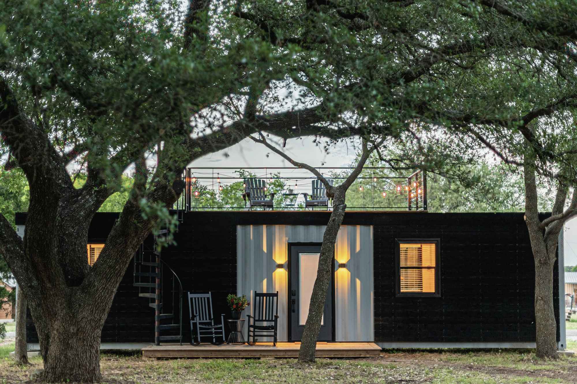 A finished dark-clad module at dusk, lit from inside, with a rooftop deck and a spiral stair, standing under live oaks.