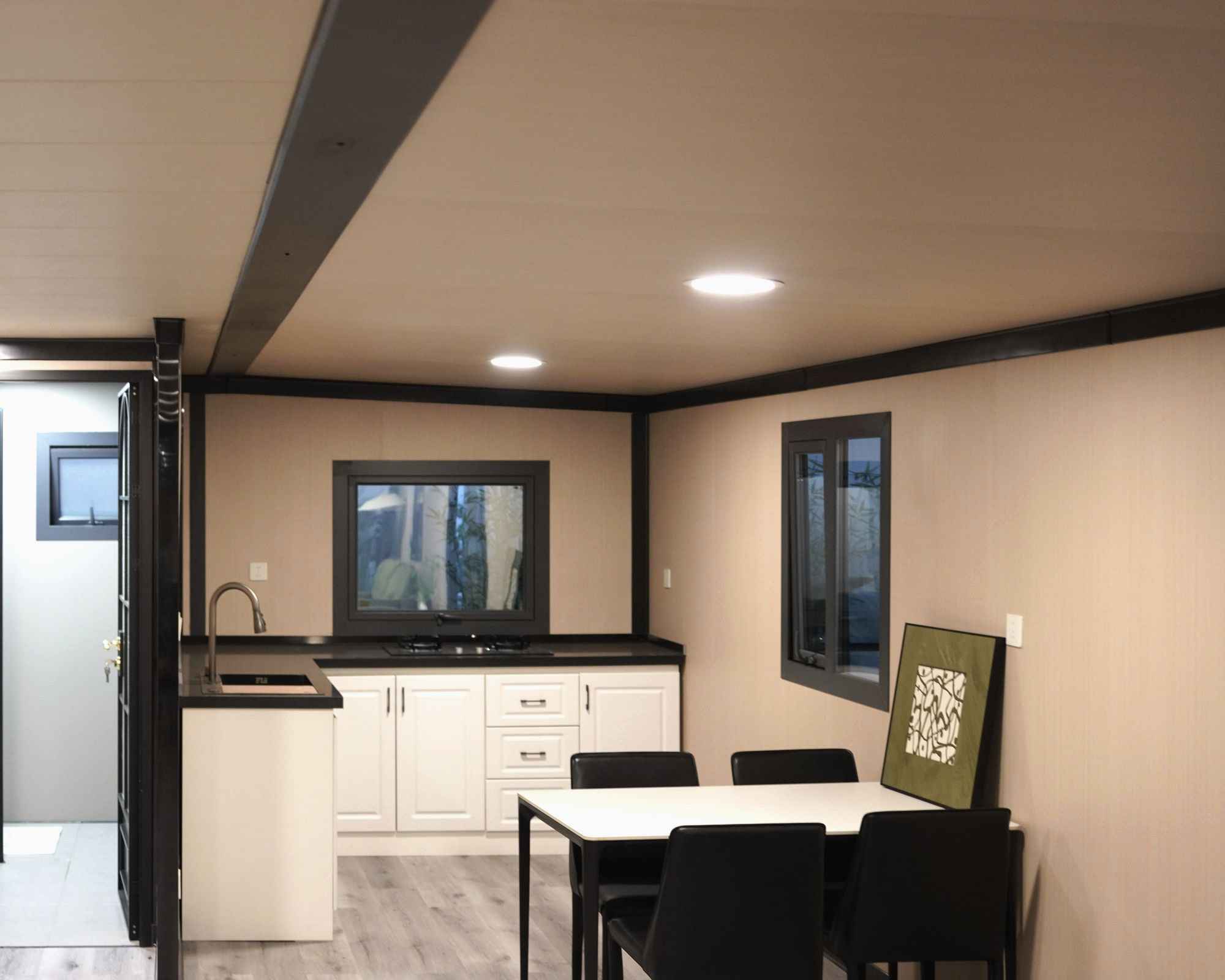 Inside a finished module: cream wall and ceiling panels, black steel frame ribs running along the wall and ceiling junctions, a kitchen along one side and a dining table at the end.