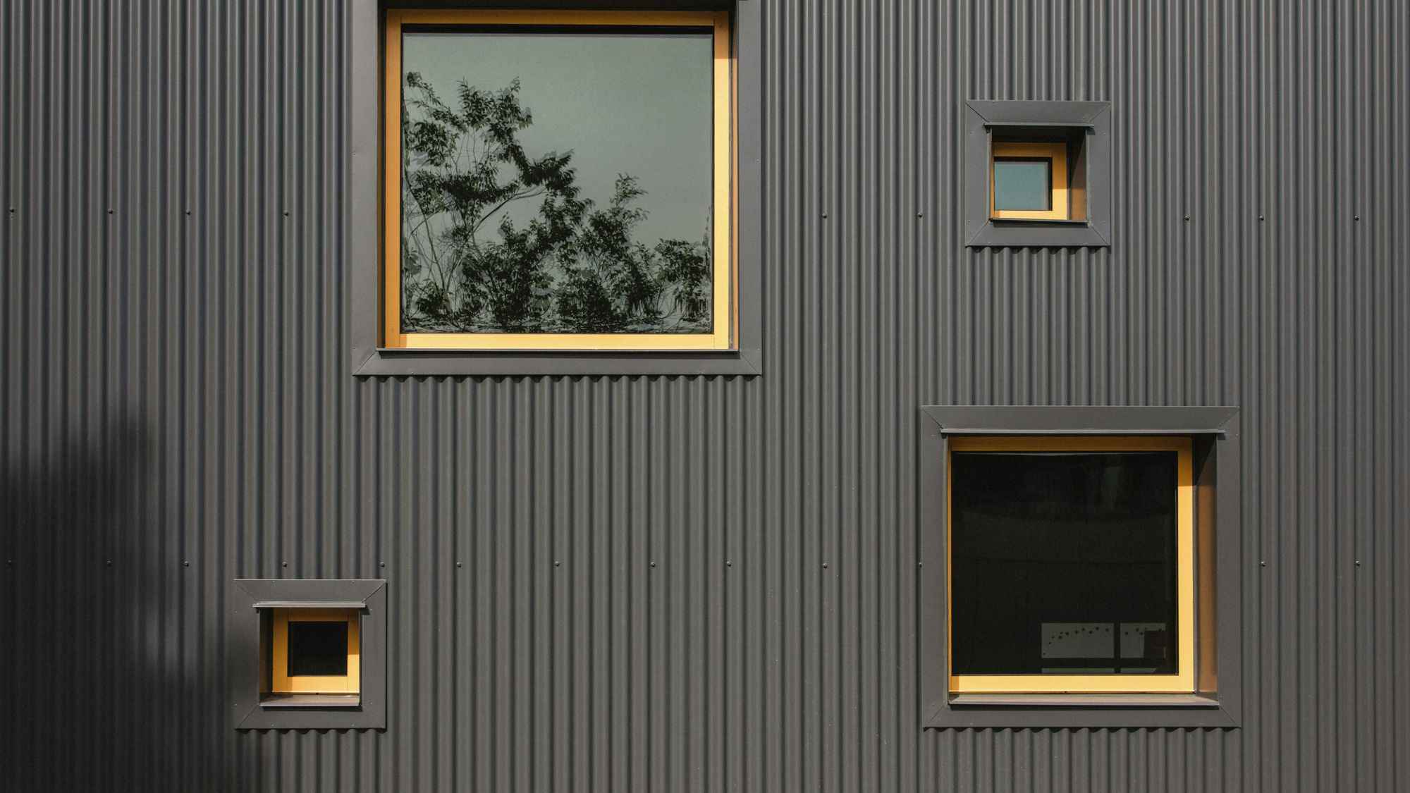Dark grey corrugated cladding with three square windows at different sizes, each set into a warm ochre reveal.